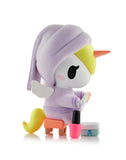 A Tokidoki Spa Day Unicorno Blind Box figurine features a kawaii Unicorno in a lavender robe and cap, seated on a stool with a nail polish palette, nail polish bottle, and small container beside it.