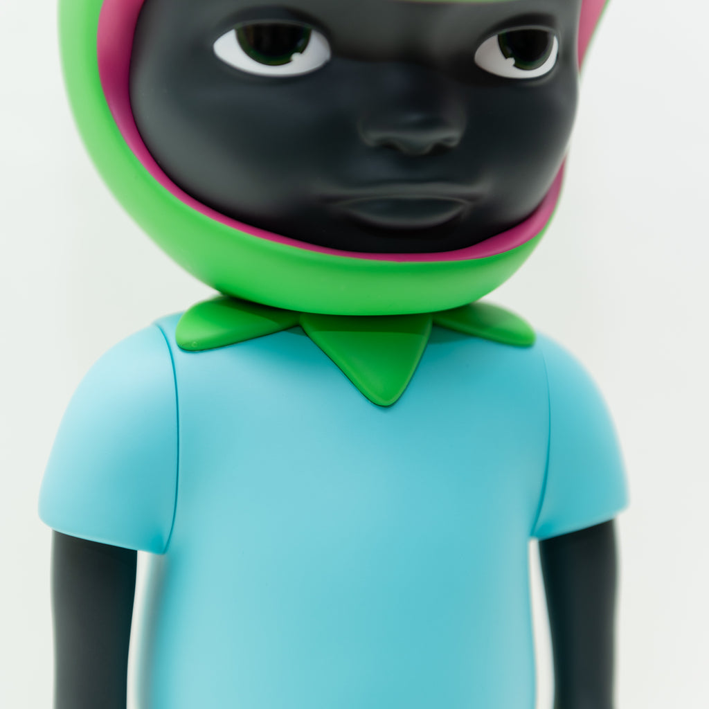 A close-up photo of a dark-skinned Phibby — OG (Green) vinyl figurine character by Hebru Brand (US), wearing a light blue shirt and a green and pink headpiece. The limited edition piece, inspired by Hebru Brantley, features a neutral expression.