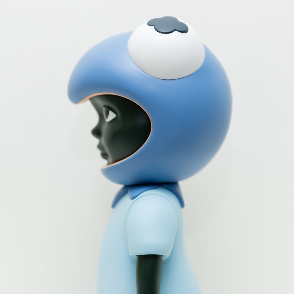 A side profile of a Hebru Brand figurine named 'Phibby — Purple' is shown with dark skin, dressed in a light blue outfit and donning a large blue helmet with a single eye on top. This limited edition piece stands against a plain white background, capturing the essence of Hebru Brantley's iconic characters.