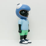 Phibby — Purple, a limited-edition toy figure by Hebru Brand (US), features dark skin and is dressed in a blue T-shirt, green shorts, white shoes, and a large blue frog-head helmet.