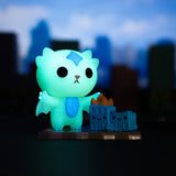 A glowing cat-like creature with dragon wings stands next to a small burning building against an out-of-focus cityscape backdrop, embodying the whimsical yet menacing charm of 100% Soft's Kaiju Kitties Blind Box.