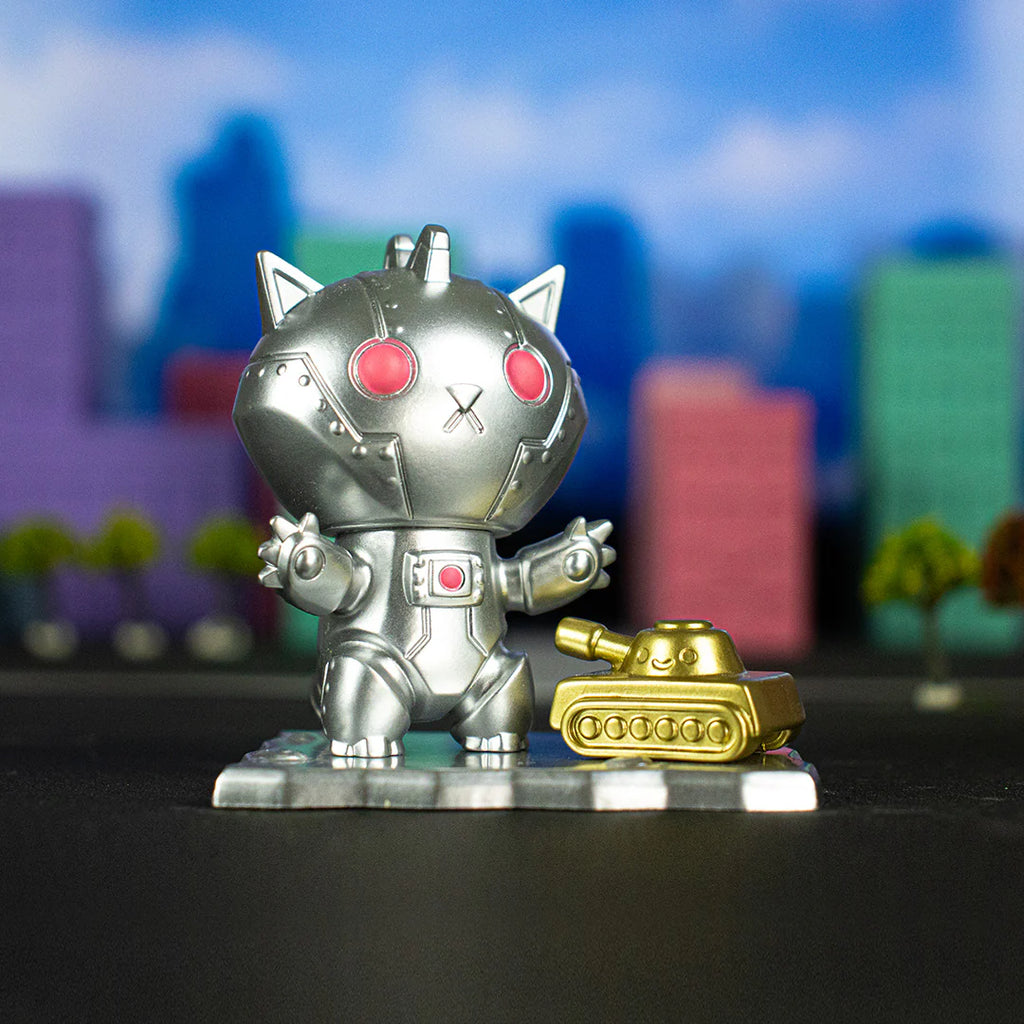 A small, silver Kaiju Kitties Blind Box figurine from 100% Soft with red eyes and a robot tank toy, looking like adorable monsters, are positioned in front of a blurry colorful cityscape background.