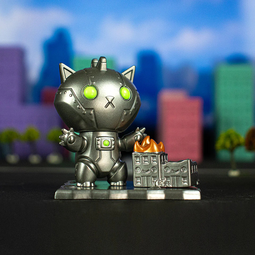 A Kaiju Kitties Blind Box by 100% Soft with green eyes stands next to a small model building on fire, conjuring scenes of adorable monsters amidst a backdrop of colorful, out-of-focus buildings and trees.