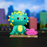 A green spiked toy creature stands on a small base next to a pink toy tank, set against a backdrop of colorful blurred buildings and trees, evoking the thrill of city destruction. The scene feels like it came straight from an exciting Kaiju Kitties Blind Box by 100% Soft.