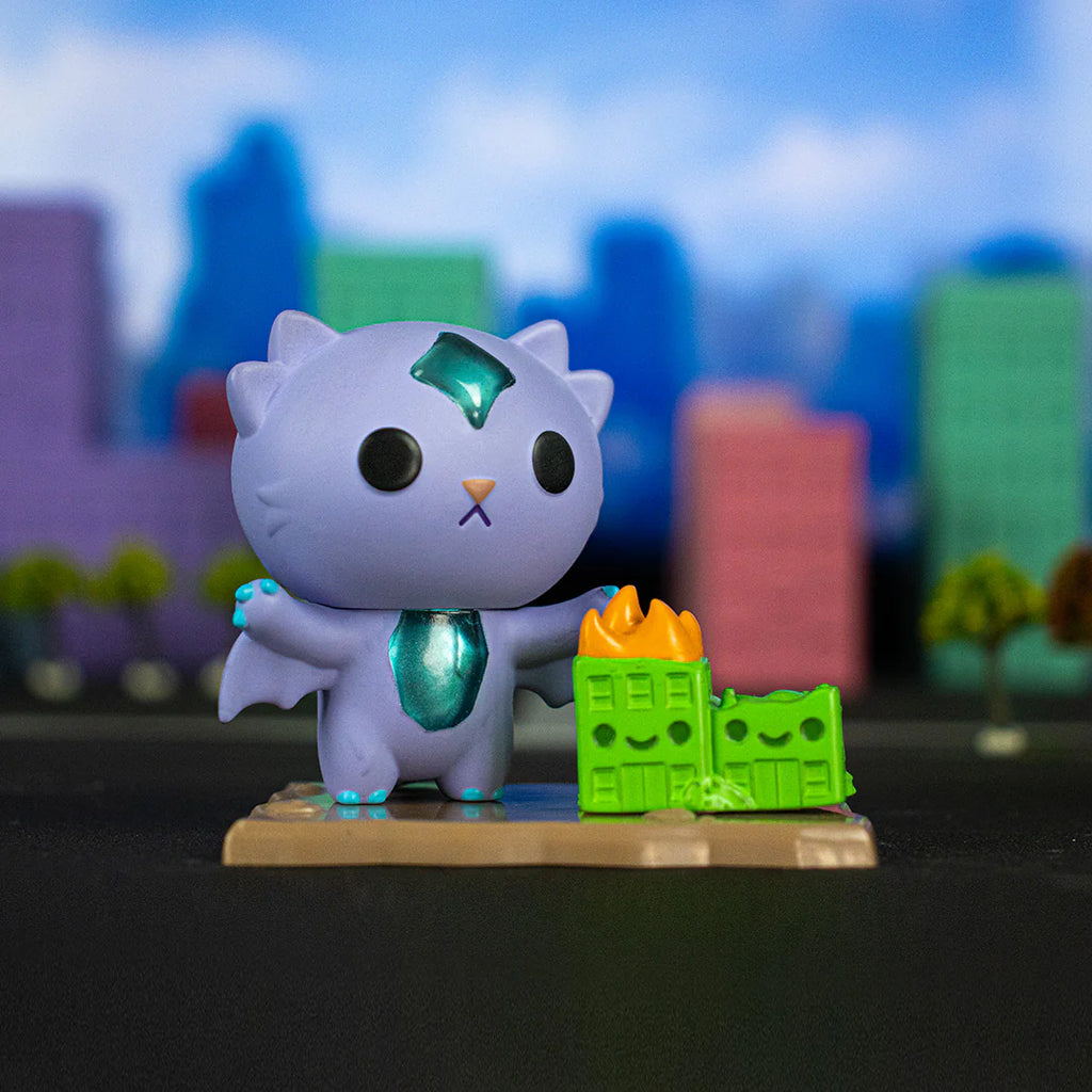 A small toy figure of a purple creature with wings and a gemstone on its forehead stands next to mini green buildings, one of which is on fire. This adorable monster looks like it came straight out of a 100% Soft Kaiju Kitties Blind Box. Colorful, blurry buildings are in the background.