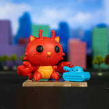 A red, cat-like figurine with tentacle-like ears holds a small blue tank toy on a stand, evoking scenes of city destruction, with a colorful, blurred cityscape in the background. It's as if plucked from the thrilling Kaiju Kitties Blind Box collection by 100% Soft featuring destructive monsters.