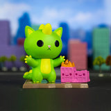 A green toy cat with a yellow belly and a dinosaur-like appearance stands on a platform with a small pink building on fire, echoing scenes of city destruction. The background shows colorful, blurry buildings. Could this be the latest addition from 100% Soft's Kaiju Kitties Blind Box series featuring destructive monsters?