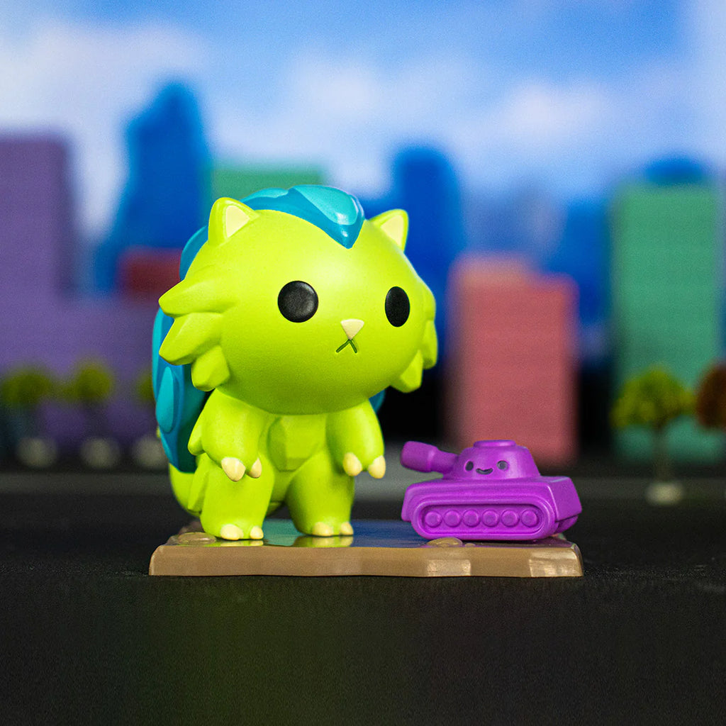 A 100% Soft Kaiju Kitties Blind Box toy figure of a green cat with blue spikes stands next to a small purple tank, reminiscent of destructive monsters, against a blurred cityscape backdrop.