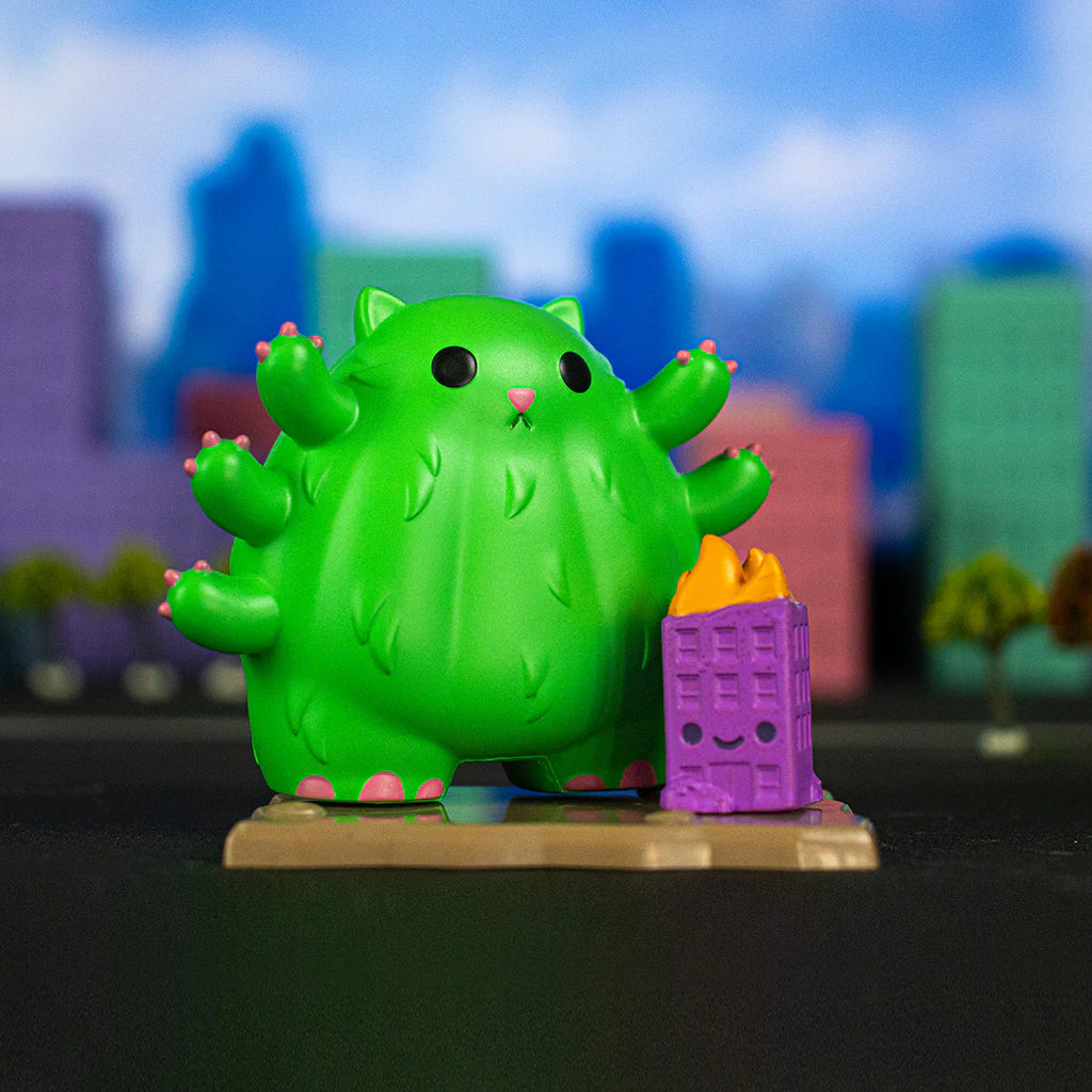 A green cactus-like cat figurine stands on a base with a small purple building on fire beside it. Evoking city destruction, the background features blurred shapes of colorful buildings against a blue sky. This Kaiju Kitties Blind Box by 100% Soft is perfect for lovers of quirky chaos.