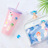 A pink tumbler adorned with Sonny Angel Stickers — Volume 1 Blind Pack by Dreams, a clear bag filled with more blind packs, and small figurines of dolls wearing animal hats sit on a light-colored surface.