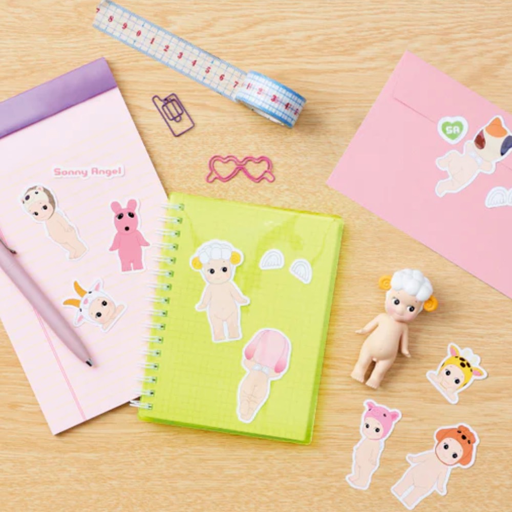 A desk with a pink notebook, a green notebook, a pink envelope, sticker packs of small dolls, a measuring tape, a pink paper clip, a pink glasses clip, and Sonny Angel dolls. There are also Dreams' water-resistant Sonny Angel Stickers — Volume 1 Blind Pack adding flair to the setup.