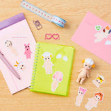 A desk with a pink notebook, a green notebook, a pink envelope, sticker packs of small dolls, a measuring tape, a pink paper clip, a pink glasses clip, and Sonny Angel dolls. There are also Dreams' water-resistant Sonny Angel Stickers — Volume 1 Blind Pack adding flair to the setup.