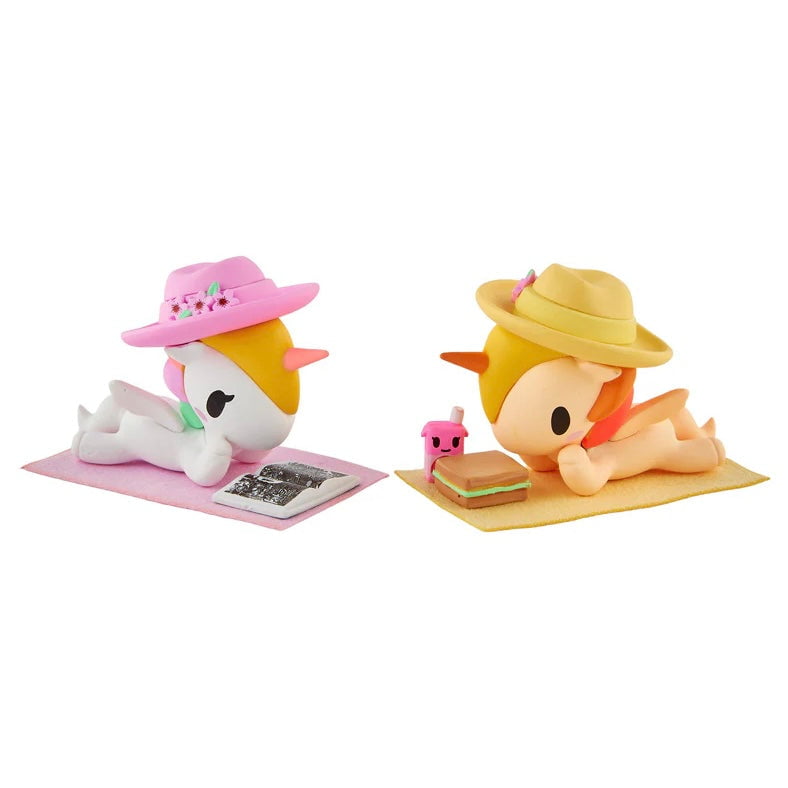 Two tokidoki Twinsies Unicorno Blind Box figures relax on mats in cartoon style—one reading a book, the other enjoying a sandwich and pink drink—adorable collectibles from tokidoki for any blind box fan.
