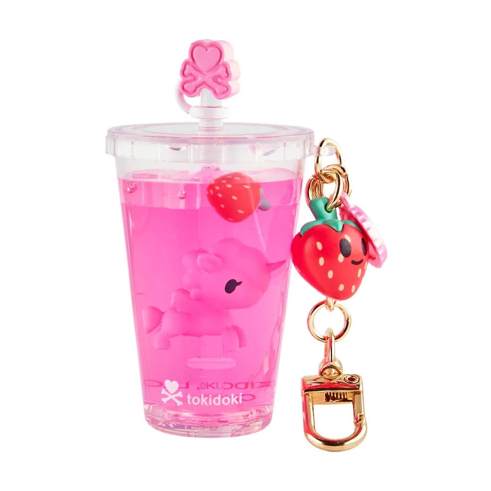 A clear cup from the tokidoki Beverage Besties Unicorno Blind Box shows pink liquid, a floating pink unicorn toy, a strawberry charm, and a gold keychain. 