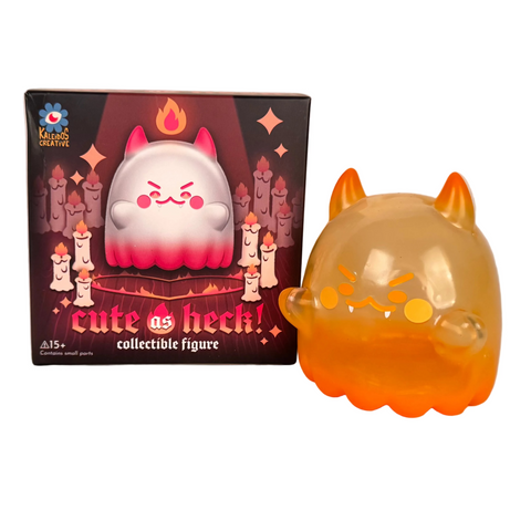 An orange and yellow cat-like Heckites figure with horns stands beside its “Cute as Heck! Blind Box” by Kaleidos Creative, with packaging featuring illustrations of the figure and candles.