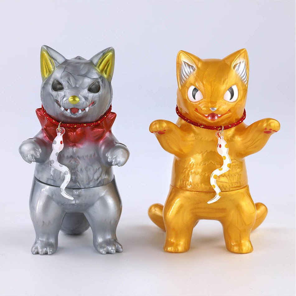 The Konatsuya 2025 Happy Set features two kaiju-inspired cat figurines: one in silver with red accents and a fish charm, and another in gold, reminiscent of Snake Cat Daruma. Both stand defiantly on a plain background. Brand: Konatsuya (JP).