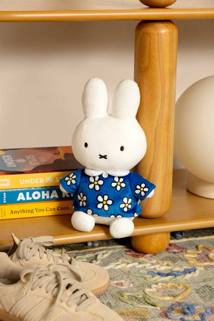 The Smoko Miffy Blue Daisy Midi Mochi Plush, a kawaii white bunny in a blue daisy dress, sits on a wooden shelf beside books, a lamp, and beige sneakers on a patterned rug.
