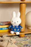 The Smoko Miffy Blue Daisy Midi Mochi Plush, a kawaii white bunny in a blue daisy dress, sits on a wooden shelf beside books, a lamp, and beige sneakers on a patterned rug.