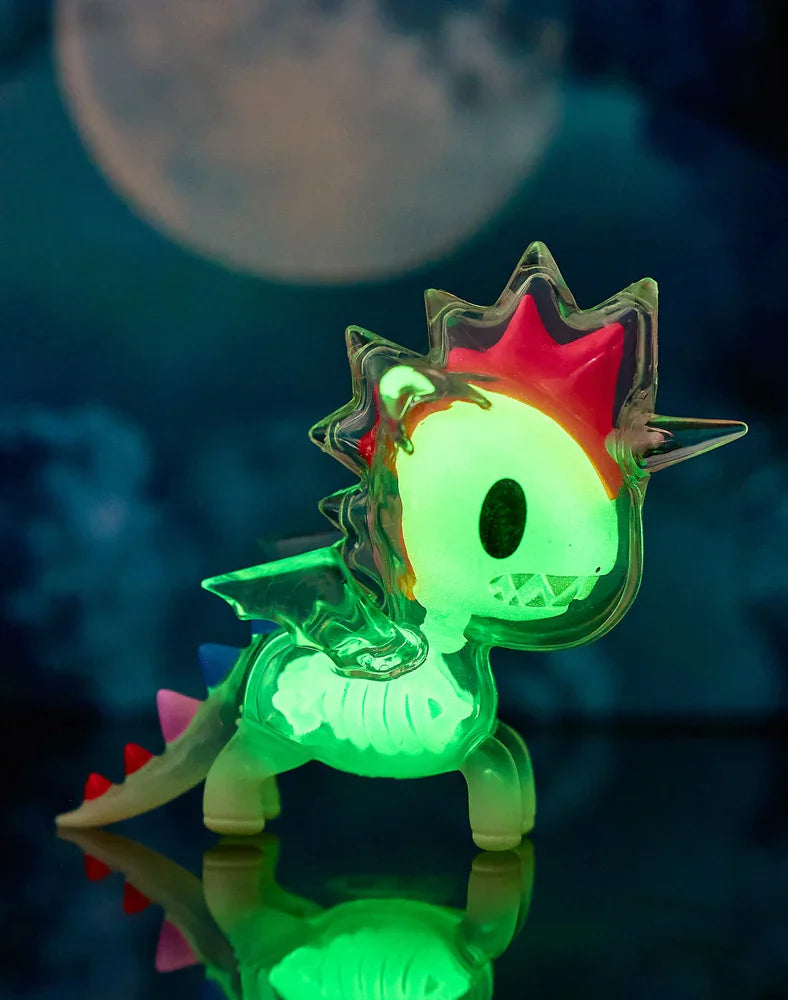 A luminous toy dragon with translucent green, pink, and blue accents stands on a reflective surface beneath a full moon and cloudy night sky, resembling the magical After Dark Skeleton Unicorno Blind Box collectible by tokidoki.