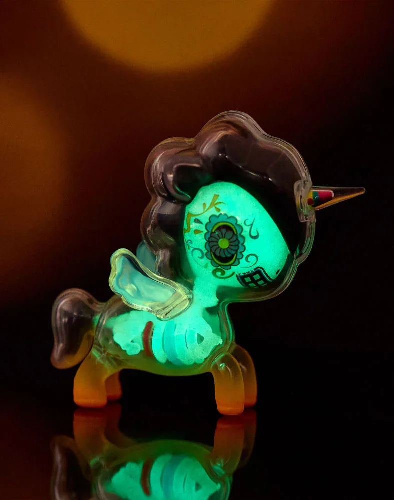 A small, glowing green tokidoki After Dark Skeleton Unicorno – Caramelo (Special Edition) stands on a shiny black surface with a blurred, warm-toned background. This collectible unicorn toy features wings, a dark mane, a decorated face, and a rainbow-colored horn. Glow-in-the-dark details make this special edition Skeleton Unicorno an enchanting centerpiece even in low light.