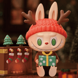 A toy bunny is holding a present in front of a Christmas tree decorated with festive costumes, featuring The Monsters - Let's Christmas Blind Box from Strangecat Toys (US).