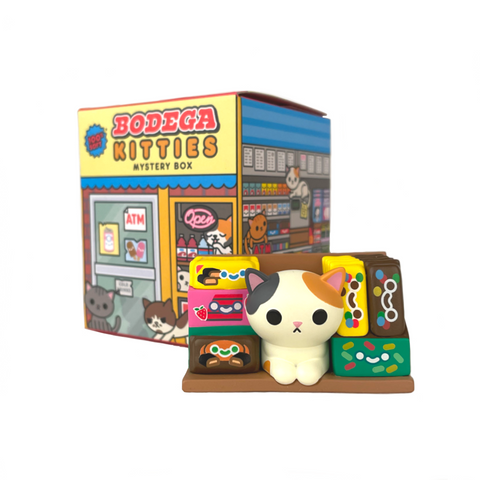 A small 100% Soft calico cat figurine is surrounded by snacks in front of a Bodega Kitties — Series 1 Blind Box, featuring cartoon cats at a bodega. Each box holds a surprise random design!.