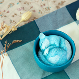 A whimsical Teacup Creatures — Cat figurine from Partner Toys features a small blue cat snuggled in a matching bowl on checkered fabric, surrounded by dried flowers.