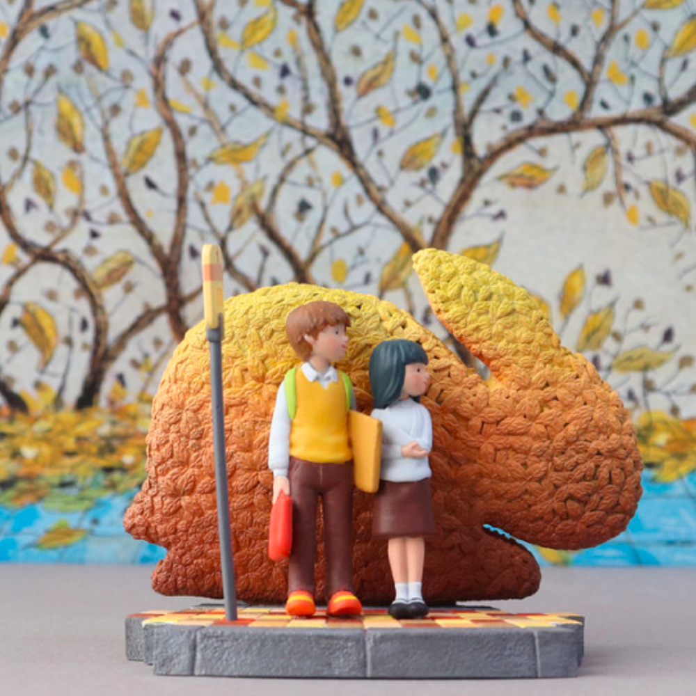 Two small figurines, a boy and a girl, from the Starry Night Blind Box by Partner Toys (TW), stand at a bus stop with an abstract rabbit sculpture in the background. The scene is set against autumn trees, like a wish come true.