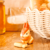 A charming Animal Bakery Blind Box figurine featuring a small rabbit with a round object sits on a wooden table next to a wicker basket filled with rabbit-shaped bread. A jar of honey from Dreams is visible in the background.