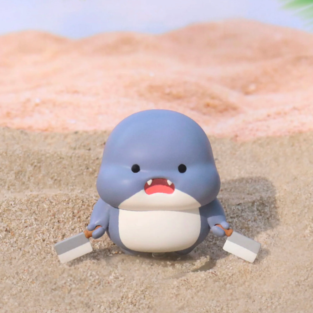 A cute cartoon Shahimi shark from the Series 2 Blind Box by Partner Toys is holding two sticks while sitting on sandy terrain.