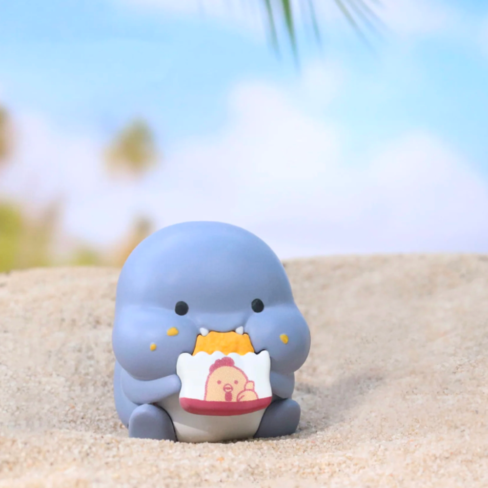 A cute and petite gray figurine from the Shahimi Shark — Series 2 Blind Box by Partner Toys is perched on sand, holding a bag of chips featuring a cartoon character, with a blurred sky and palm trees in the background. This charming piece is part of the delightful Shahimi collection, resembling the joy of discovering a surprise in every blind box.