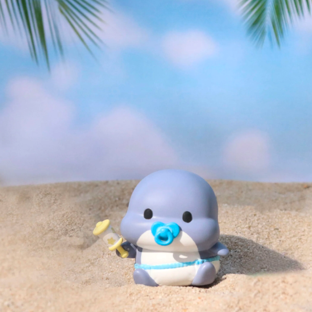 A small Shahimi Shark from Partner Toys, adorned in gray and with a pacifier, lounges on a sandy beach under palm fronds, holding a yellow syringe toy. Nearby, a Series 2 Blind Box is nestled in the sand, promising surprises within, all set against a vibrant blue sky.