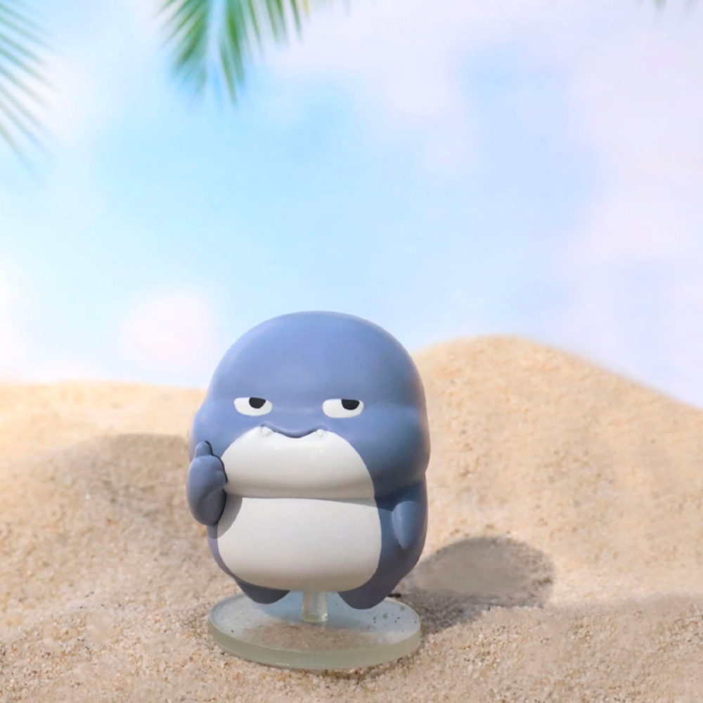 A small, round toy figure from the Shahimi Shark — Series 2 Blind Box by Partner Toys stands on sandy terrain with palm leaves overhead and a blue sky backdrop.