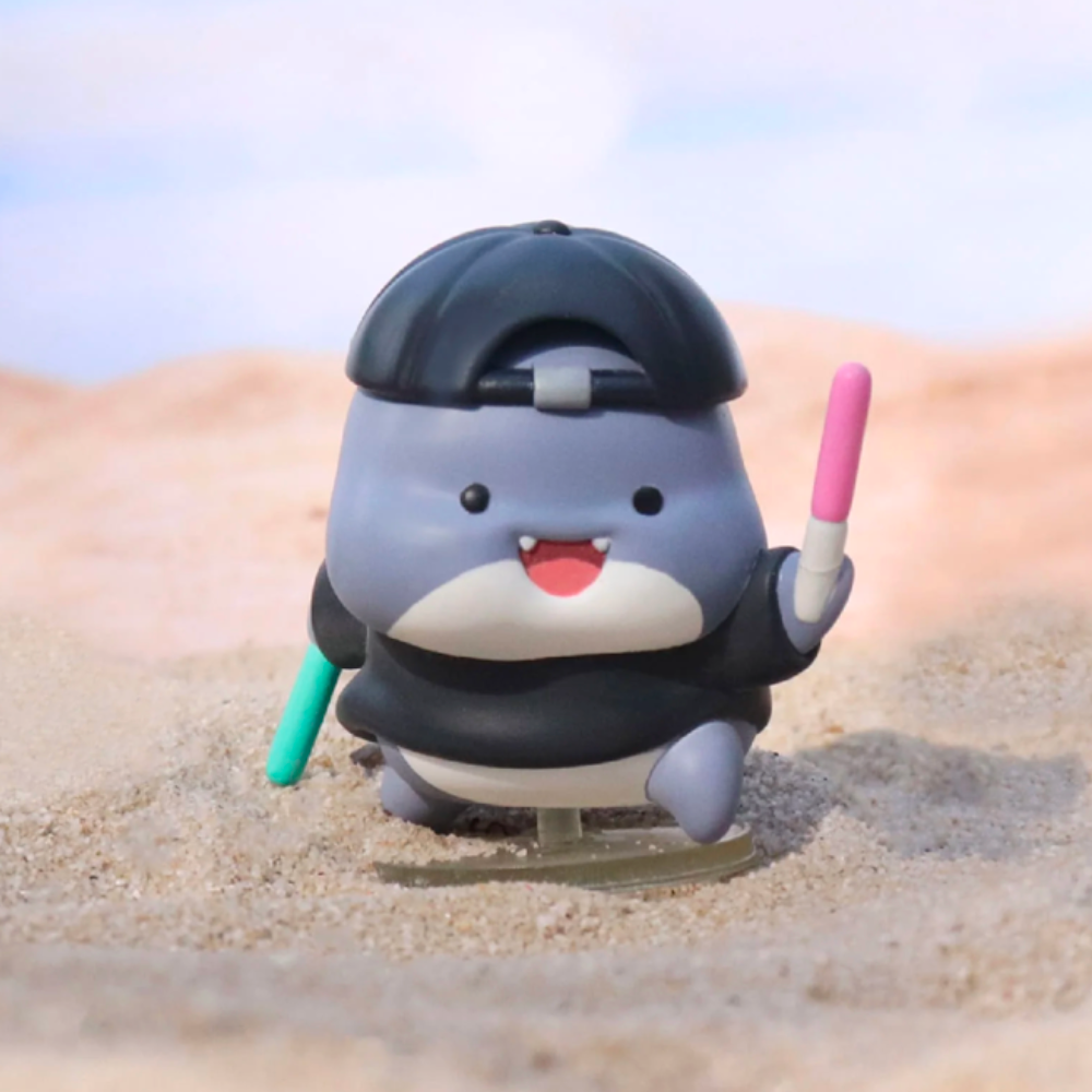 A charming gray and white Shahimi Shark figurine by Partner Toys, from the Series 2 Blind Box, boasts a black cap and holds two colorful sticks, sitting on sand like a hidden treasure.