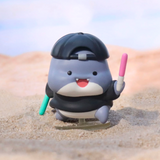 A charming gray and white Shahimi Shark figurine by Partner Toys, from the Series 2 Blind Box, boasts a black cap and holds two colorful sticks, sitting on sand like a hidden treasure.