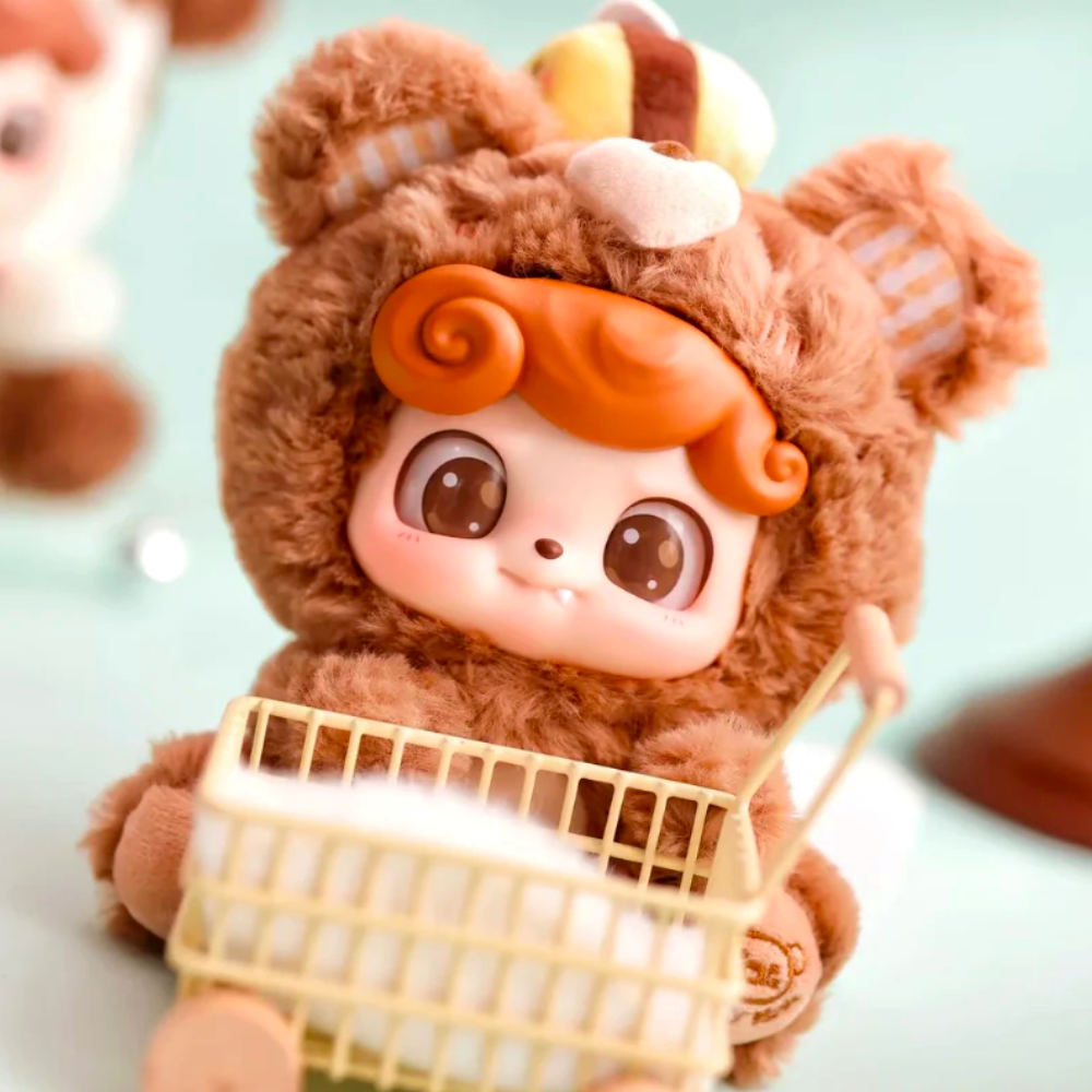 A cute baby doll from the Jotoys Q.Kid Animal PreSchool Plush Blind Box, featuring orange hair in an animal costume, is next to a small shopping cart on a light green background.