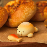 Experience the whimsical delight of our Dreams' Animal Bakery Series 2 Blind Box. Each adorably crafted seal-shaped bread roll, complete with a tiny fish-shaped piece, sits on brown parchment paper, ready to surprise you with its charming animal-inspired forms amidst other baked delights.