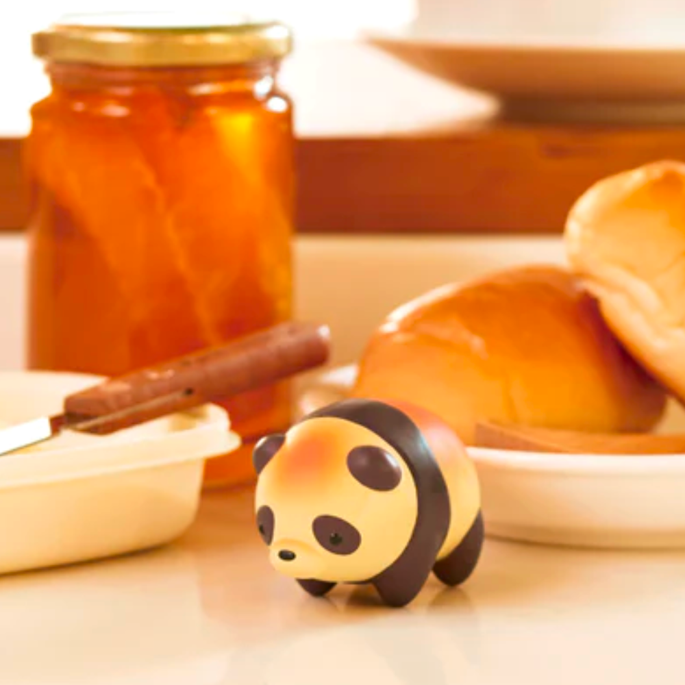 A miniature figurine from the Dreams Animal Bakery Blind Box is placed on a kitchen counter in front of a jar of honey, a butter knife, and a dish with rabbit-shaped bread rolls.