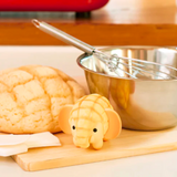 A metal mixing bowl and whisk are on a wooden cutting board next to a loaf of bread and an elephant-shaped bread from the Dreams Animal Bakery Blind Box.