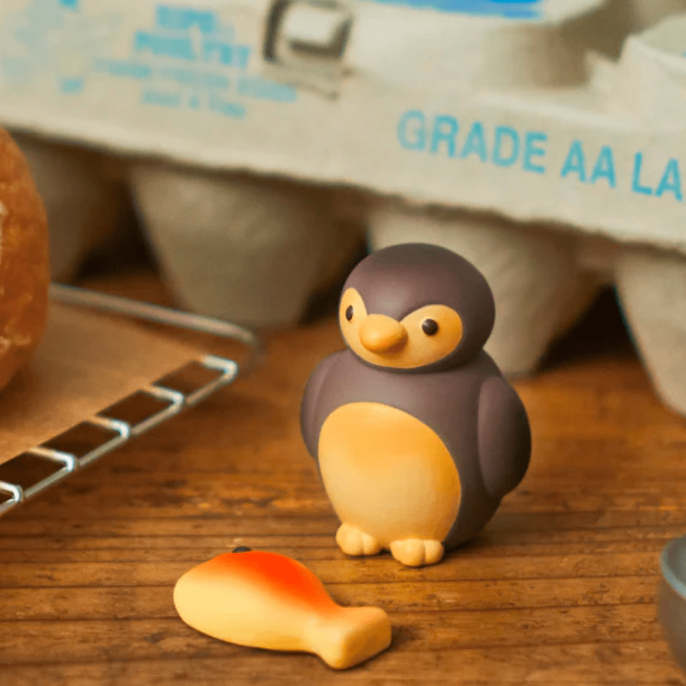 A small toy penguin and toy fish rest on a wooden surface with a carton of eggs in the background, while Dreams' 