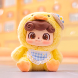 A Q.Kid Animal PreSchool Plush Blind Box doll by Jotoys, dressed in a yellow duck costume with a gingham bib and small hat, sits on a smooth surface against a blurred orange backdrop.