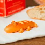 An adorable orange, crocodile-shaped toy resembling bread from Dreams' Animal Bakery Series 2 Blind Box sits on a white cloth, with real bread and a red carton in the background, creating a delightful scene filled with charming surprises.