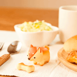 A small toy dog and a bone-shaped object from the Dreams Animal Bakery Blind Box are on a table with a bowl of salad, a cup, a fork, a spoon, and a plate of rabbit-shaped bread pastries.