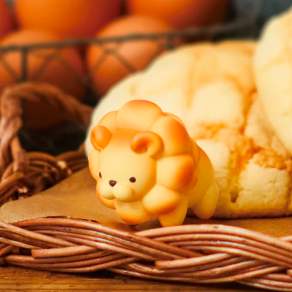 A small, cute bread-shaped lion toy from Dreams' Animal Bakery Series 2 Blind Box sits on a wicker basket beside a large round loaf, with blurred oranges in the background.