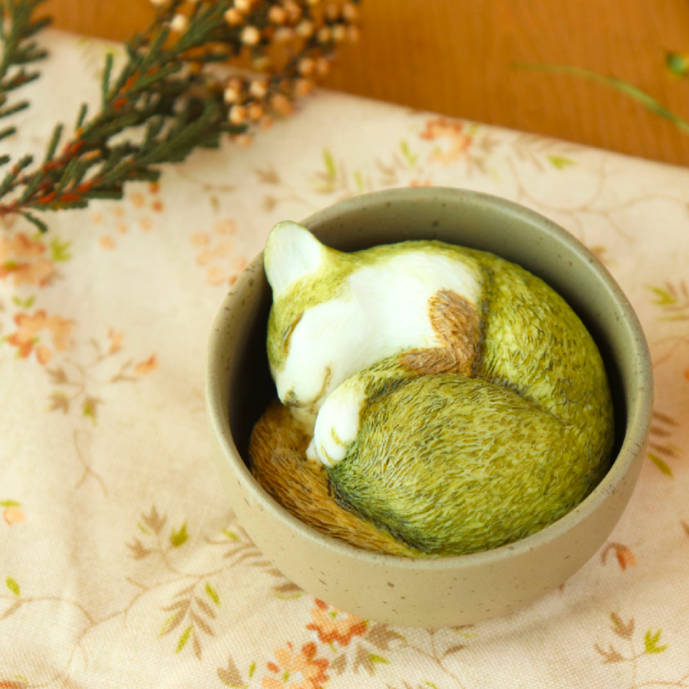 A Teacup Creatures — Cat Blind Box by Partner Toys features a realistic cat figurine curled up asleep in a small brown bowl on floral-patterned cloth with greenery, reminiscent of a surprise find.