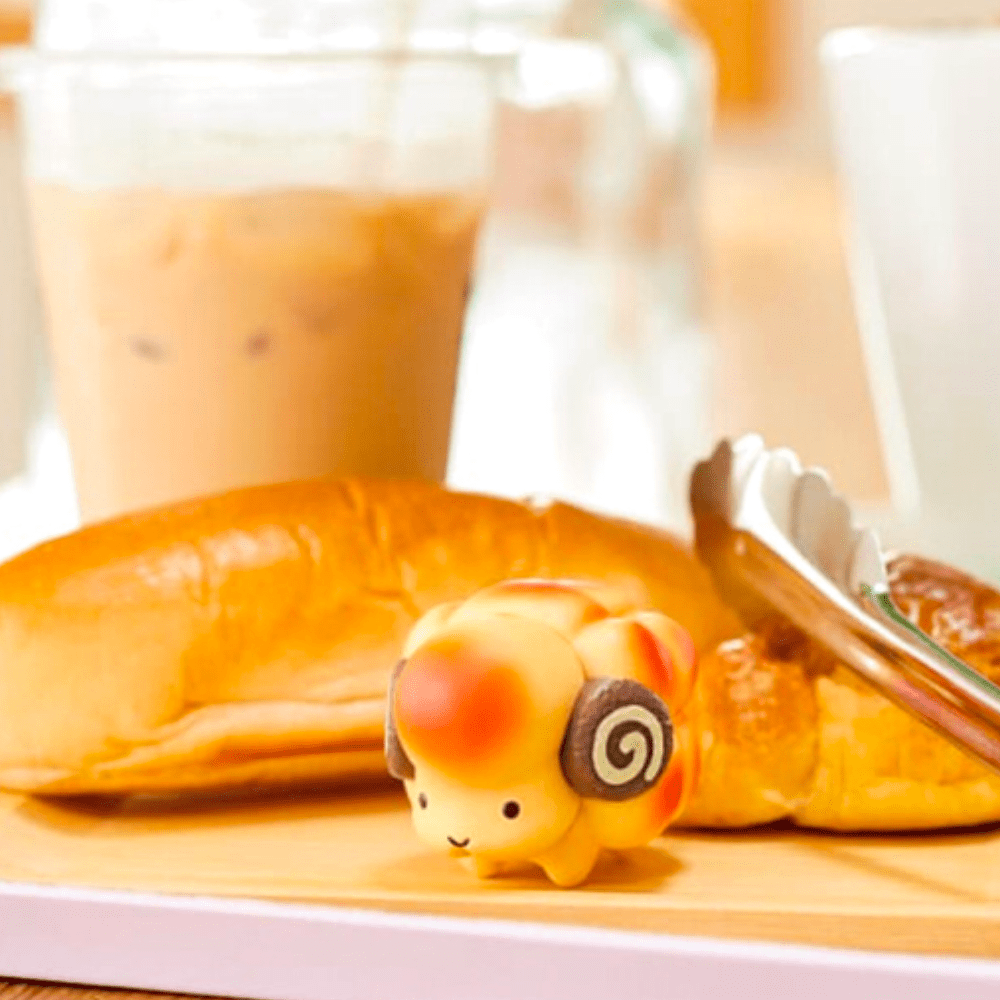 A Dreams Animal Bakery Blind Box featuring a toy sheep is placed in front of rabbit-shaped bread rolls and tongs on a tray. A glass of iced coffee can be seen in the background.