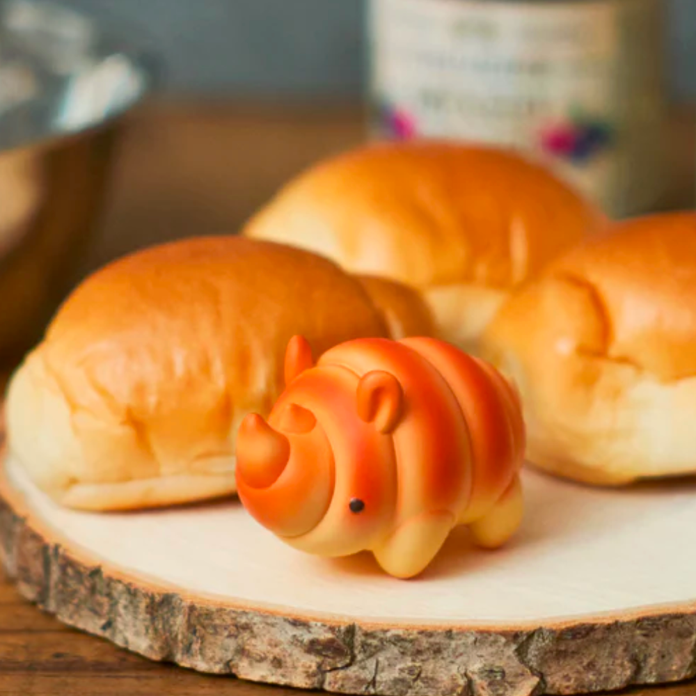 A charming orange rhino figurine from the Dreams Animal Bakery Series 2 Blind Box sits adorably on a wooden board, surrounded by three cute bread rolls that look like they're part of a blind box collection.