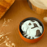 A Teacup Creatures—Cat Blind Box by Partner Toys features a small clay model of a sleeping white cat with brown spots, curled inside a terracotta pot, resting on a wooden surface adorned with dried flowers.