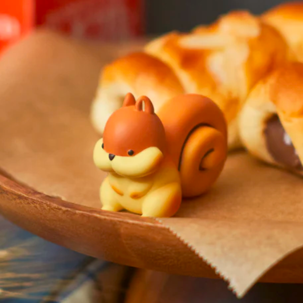 A cute squirrel figurine from the Animal Bakery Series 2 Blind Box by Dreams sits on a brown plate near a croissant. Its brown and yellow colors and charming spiral tail give it an adorable, bread-like appearance.