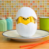 A Gudetama plush from Gund, resembling a cracked egg sits on a plate, playing peek-a-boo with sleepy eyes. Nearby, a cup, salt and pepper shakers, and chopsticks rest against vibrant tiles.